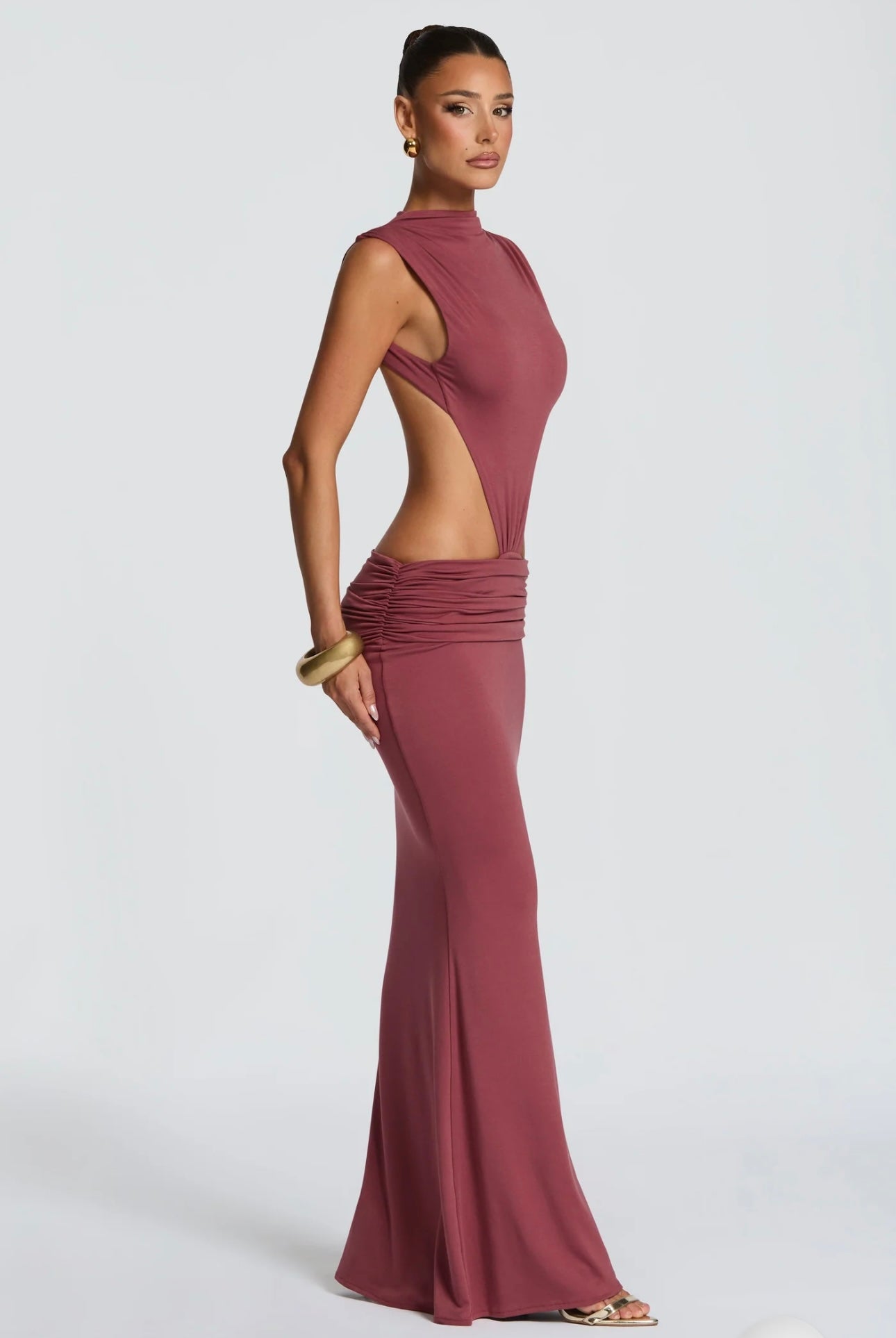 Abito Sahara maxi aderente in elegante rosa terracotta con design cut-out laterale che scolpisce la silhouette. Il collo alto crea un raffinato contrasto con le ampie aperture sui fianchi e la schiena scoperta, per un effetto sensuale ma sofisticato.