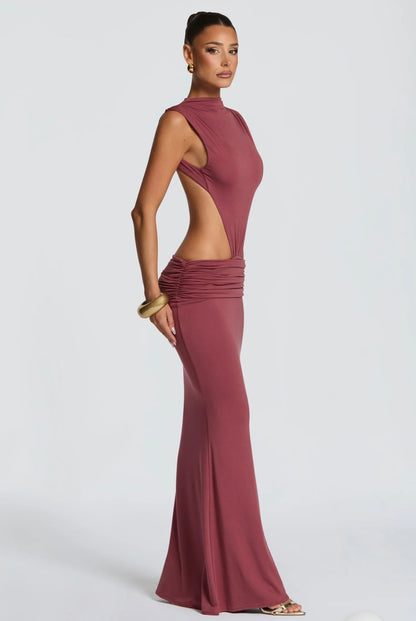 Abito Sahara maxi aderente in elegante rosa terracotta con design cut-out laterale che scolpisce la silhouette. Il collo alto crea un raffinato contrasto con le ampie aperture sui fianchi e la schiena scoperta, per un effetto sensuale ma sofisticato.