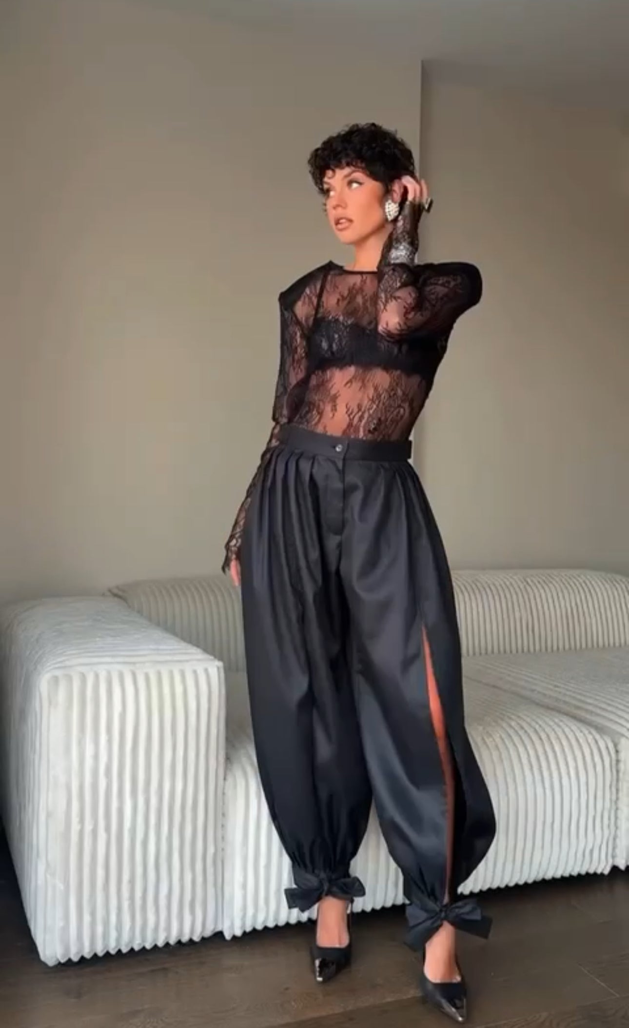 Outfit Gabriella elegante e sensuale composto da maglia in pizzo trasparente e pantalone nero a vita alta. Il pantalone presenta una linea fluida con spacco laterale e dettagli con fiocchi alla caviglia, per un look sofisticato e di grande impatto.