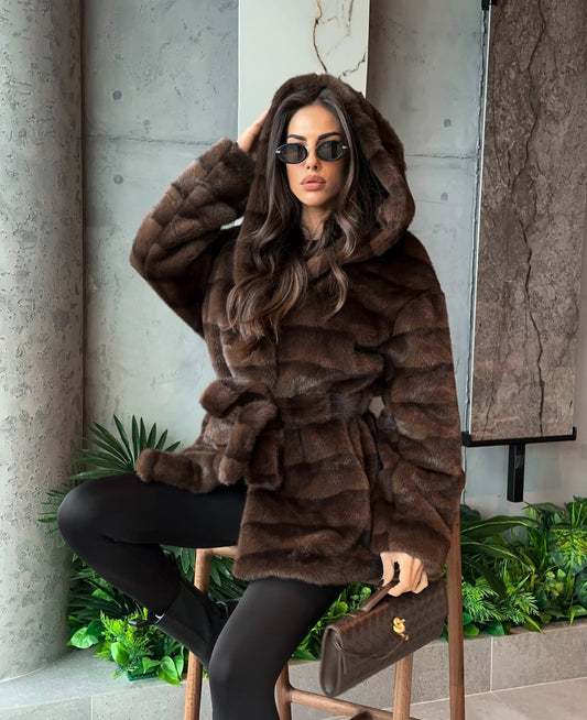 Pelliccia GEMMA Realizzato in pelliccia di visone ecologico di alta qualità, presenta una texture morbida e avvolgente con lavorazione a fasce orizzontali. Il design è arricchito da cappuccio e cintura in coordinato