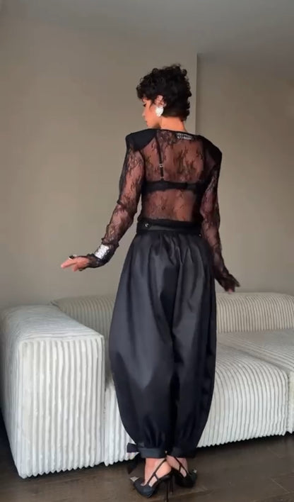 Outfit Gabriella elegante e sensuale composto da maglia in pizzo trasparente e pantalone nero a vita alta. Il pantalone presenta una linea fluida con spacco laterale e dettagli con fiocchi alla caviglia, per un look sofisticato e di grande impatto.