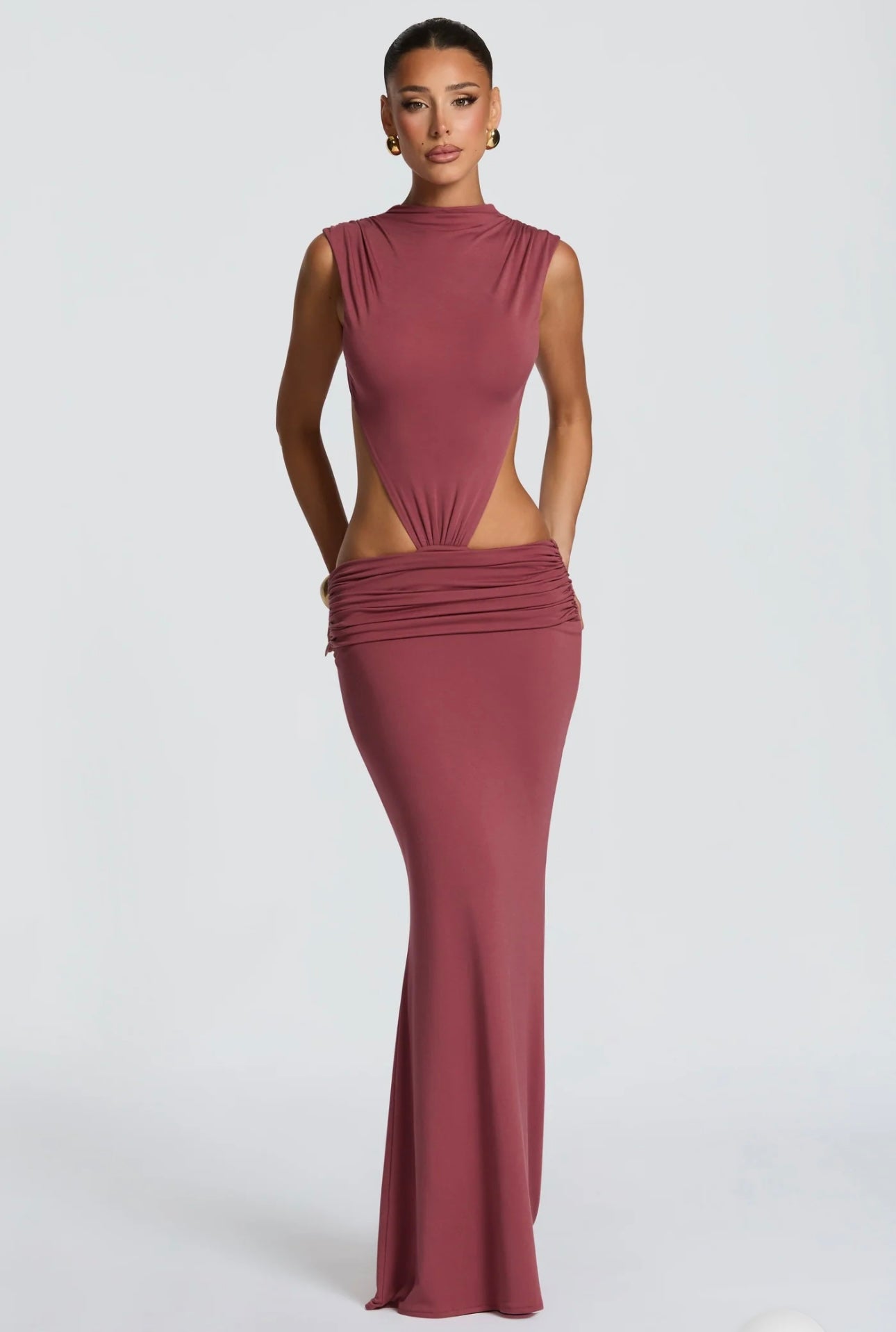 Abito Sahara maxi aderente in elegante rosa terracotta con design cut-out laterale che scolpisce la silhouette. Il collo alto crea un raffinato contrasto con le ampie aperture sui fianchi e la schiena scoperta, per un effetto sensuale ma sofisticato.